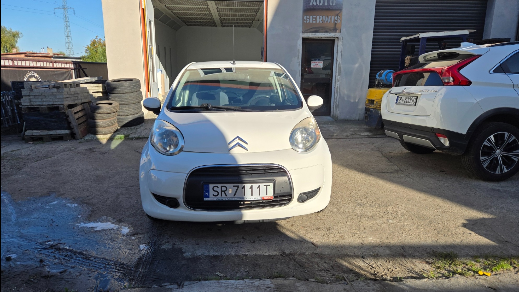 Biały Citroen C1 przed warsztatem samochodowym z otwartą bramą. Widoczne opony i palety po lewej stronie. Drugi samochód częściowo widoczny z tyłu po prawej.