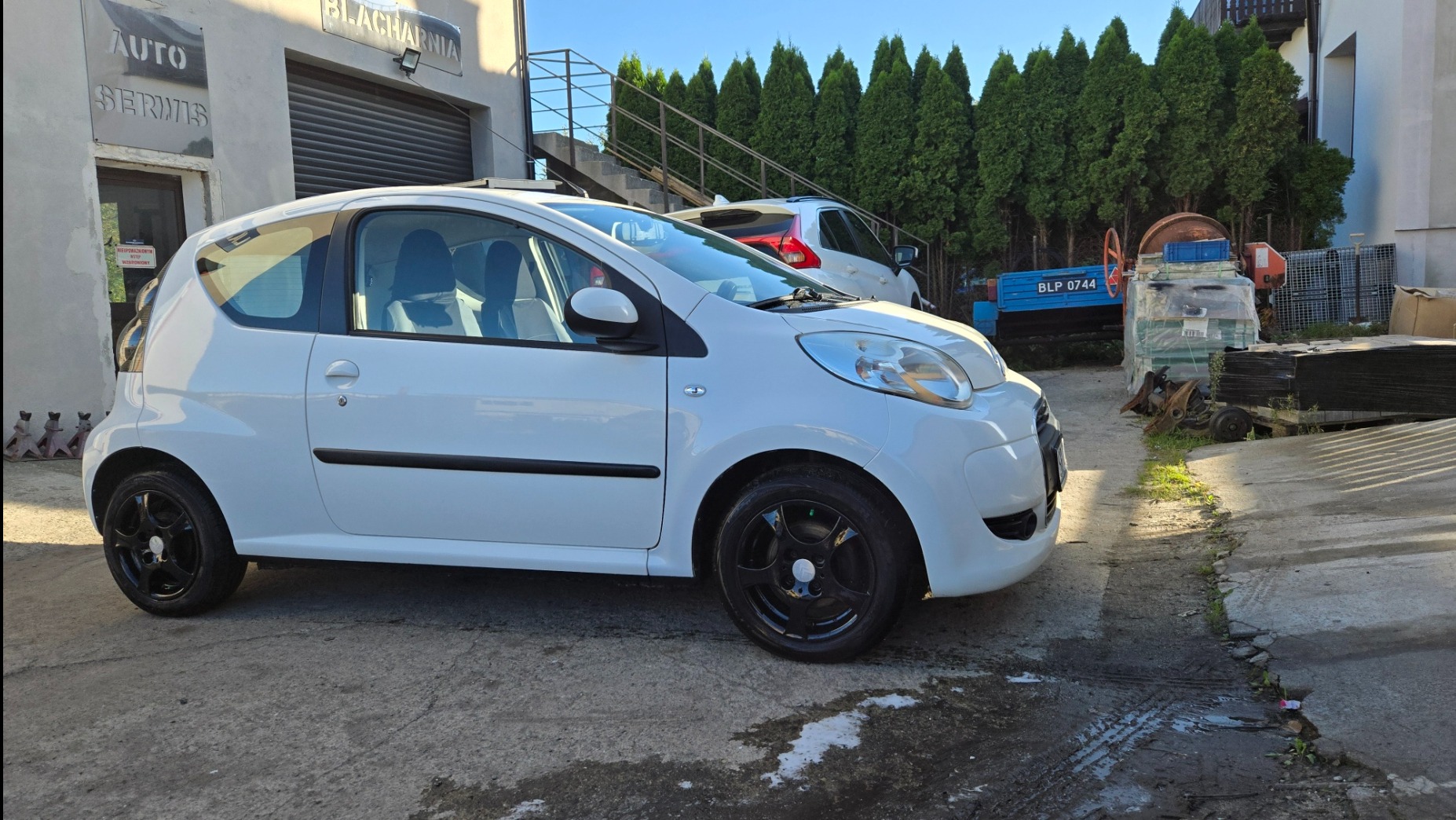 Biały samochód przed budynkiem z napisem 'Blacharnia'. Czarny felgi, jasne wnętrze. Ujęcie z boku, widoczne inne pojazdy w tle.