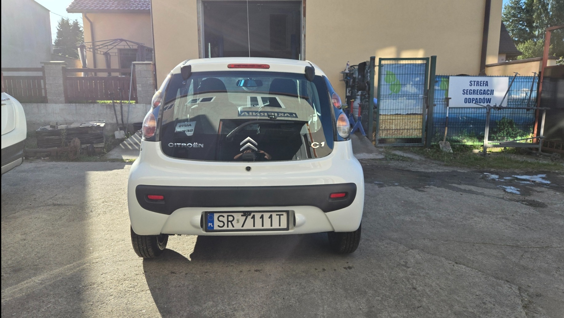 Tył białego Citroena C1 z widoczną tablicą rejestracyjną SR 7111T, zaparkowanego przed garażem i ogrodzeniem z napisem 'Strefa segregacji odpadów'.