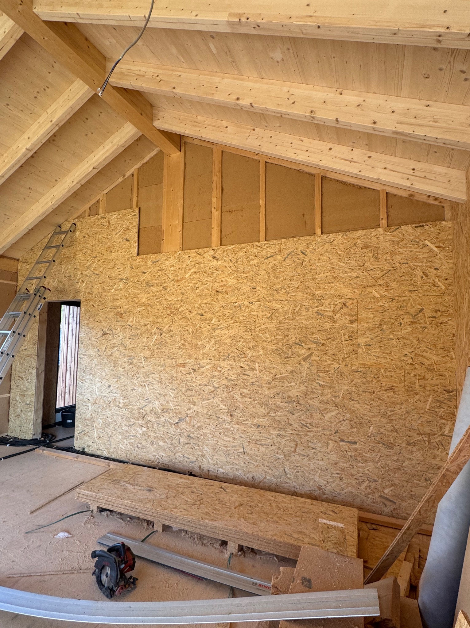 Wnętrze w trakcie wykańczania płytami OSB: ściany, drewniany szkielet dachu, drabina oparta o ścianę, narzędzia i materiały budowlane na podłodze.