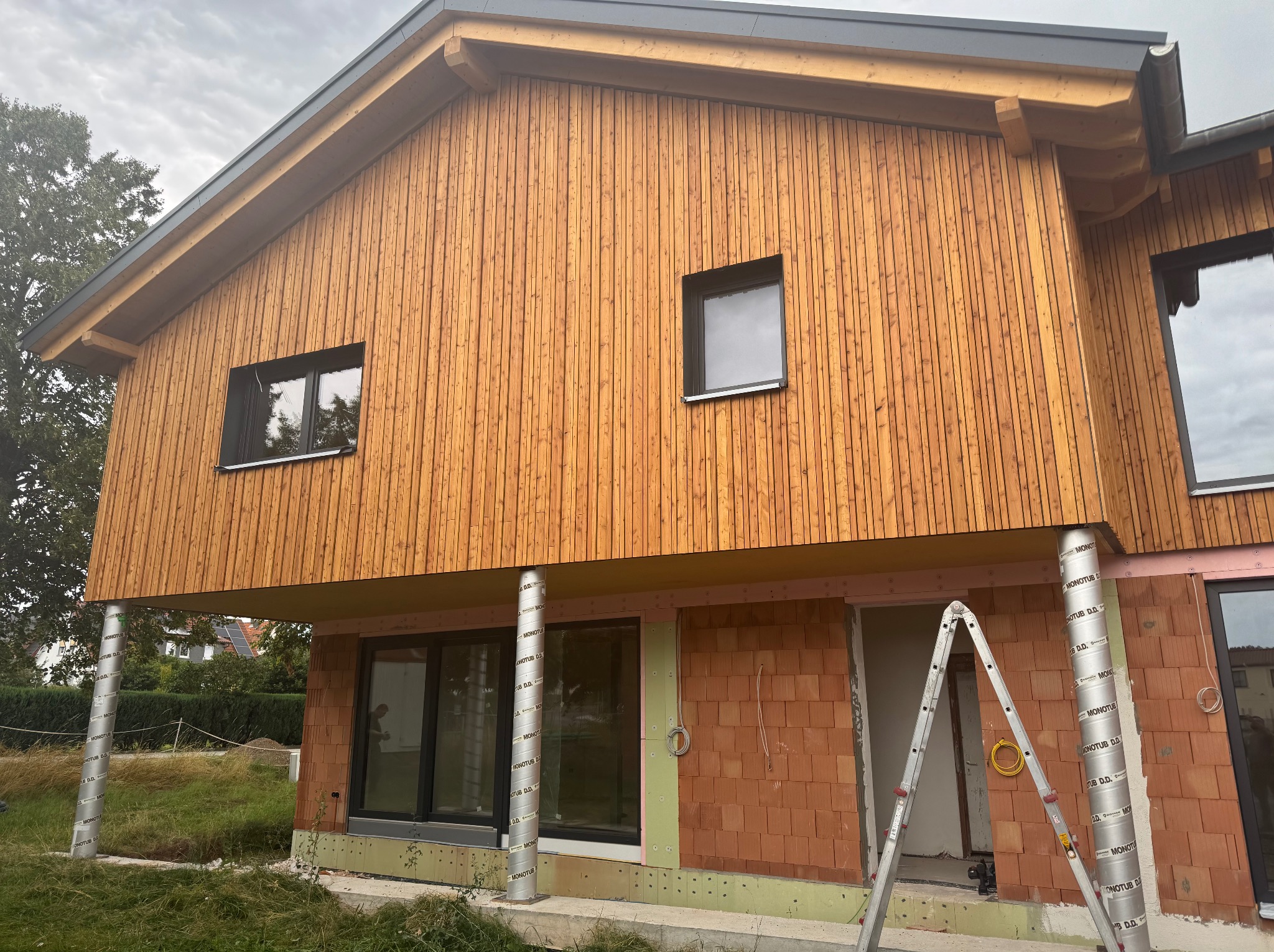 Dom w trakcie budowy: drewniana elewacja na piętrze, ceglana ściana na parterze, widoczne okna i drabina. Budynek na słupach, stan surowy.