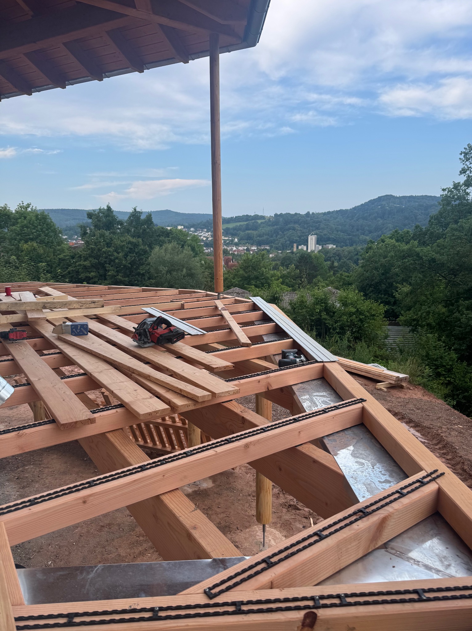 Konstrukcja drewniana dachu w trakcie budowy, widok z góry na elementy szkieletu, narzędzia pracy i panoramę miasta w tle. Szczegółowe połączenia i materiały budowlane.