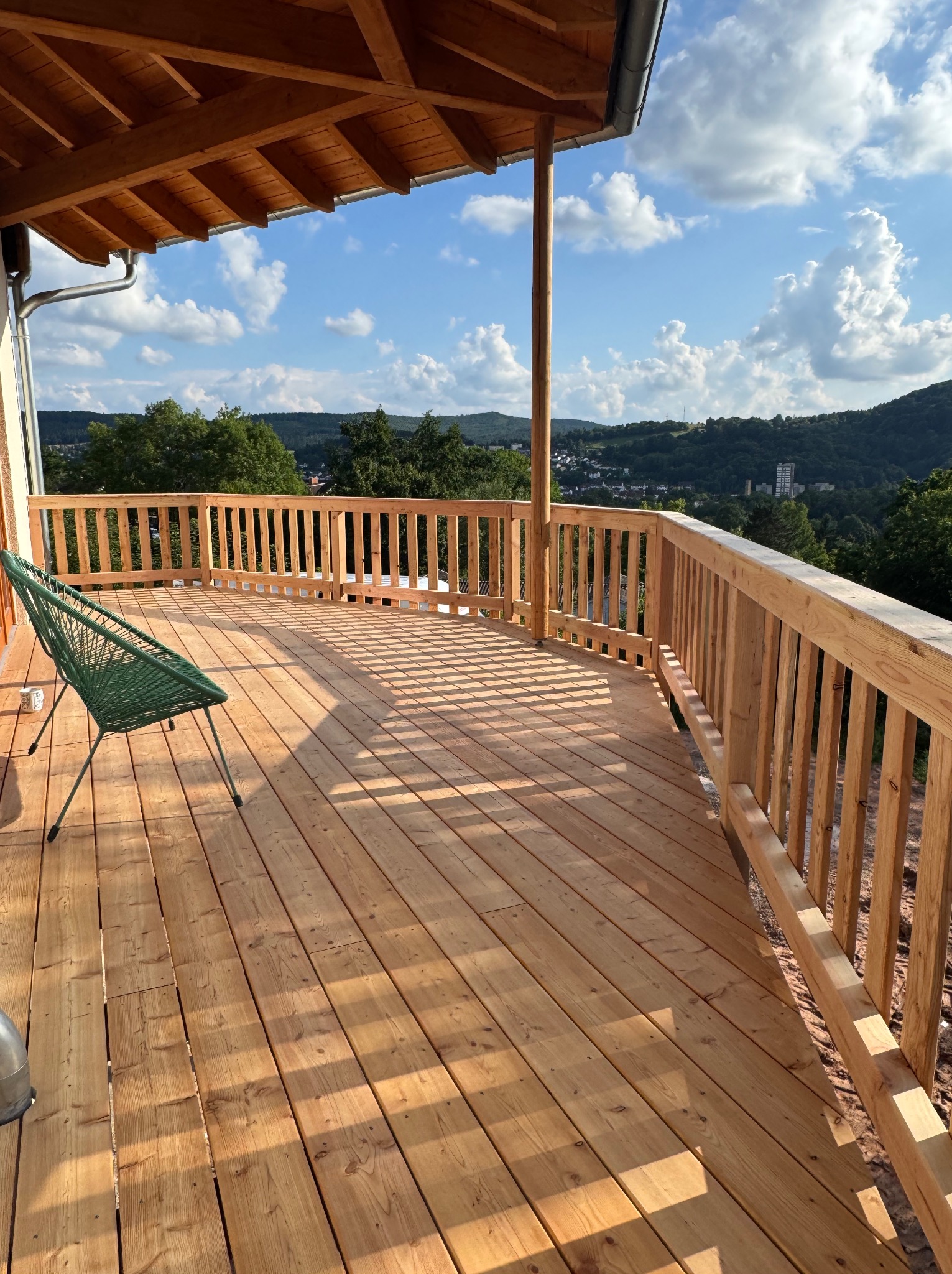 Nowy drewniany taras z widokiem na góry, oświetlony słońcem. W tle zielone wzgórza i błękitne niebo z chmurami. Na tarasie stoi zielone krzesło.