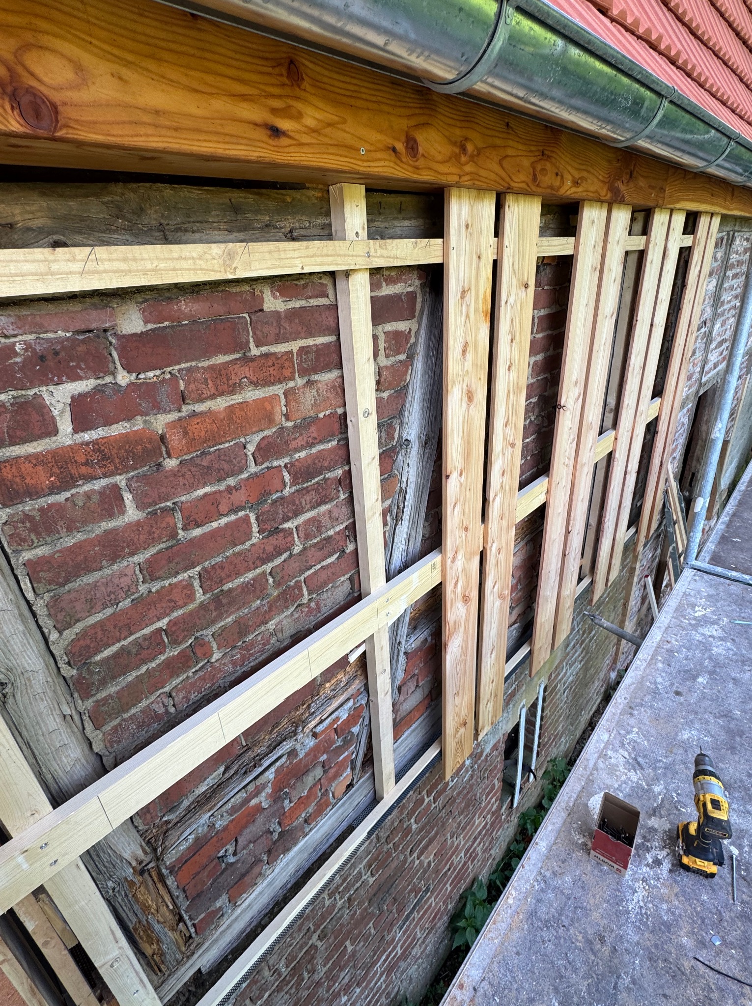 Ceglana ściana z drewnianą konstrukcją i listwami, widok z rusztowania. W tle rynna i dachówka. W pobliżu wkrętarka i pudełko z wkrętami.