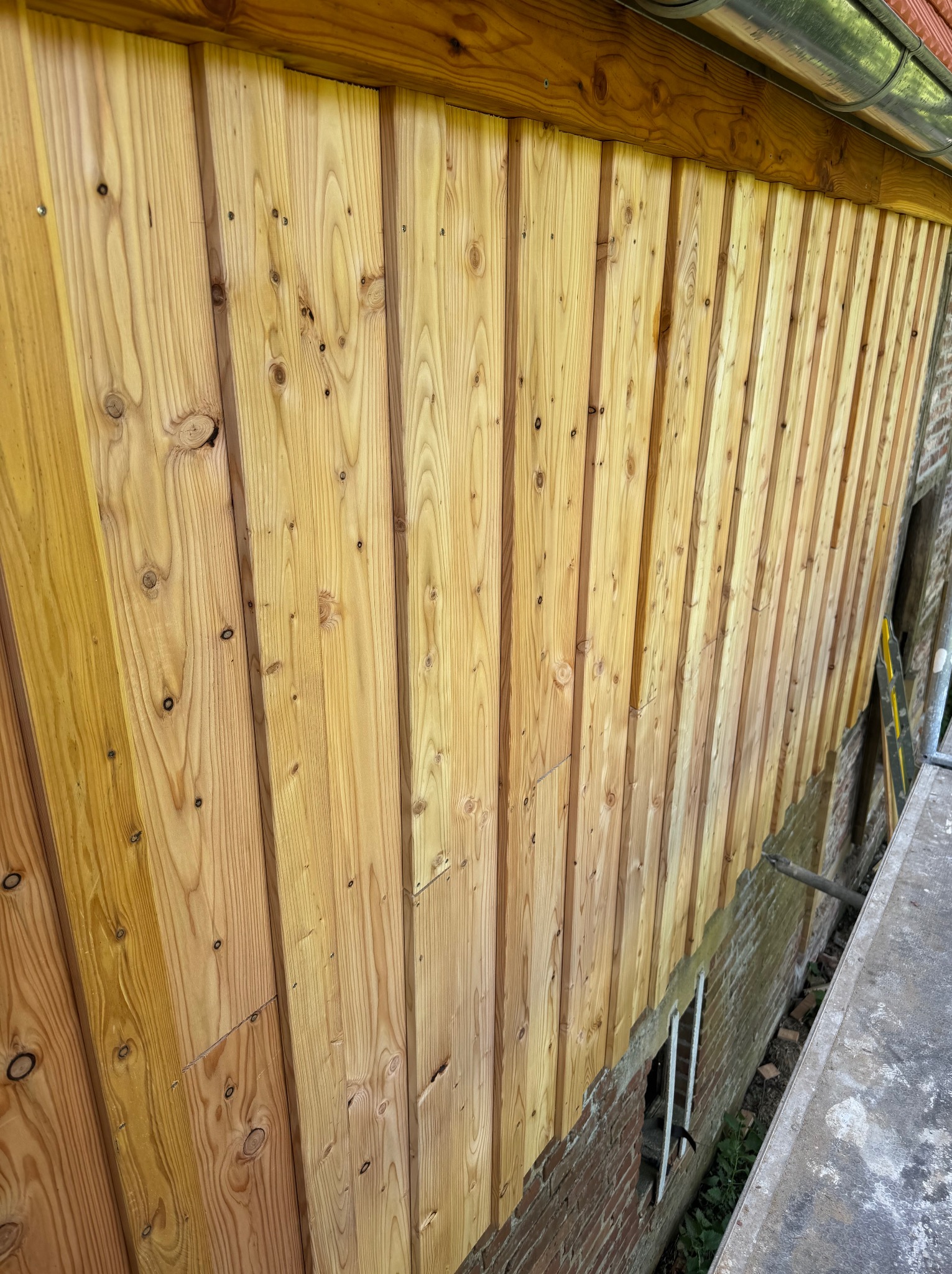 Ściana obita jasnym drewnem, pionowe deski z widocznymi słojami i sękami, fragment muru z cegły, praca w toku, widoczne elementy rusztowania.