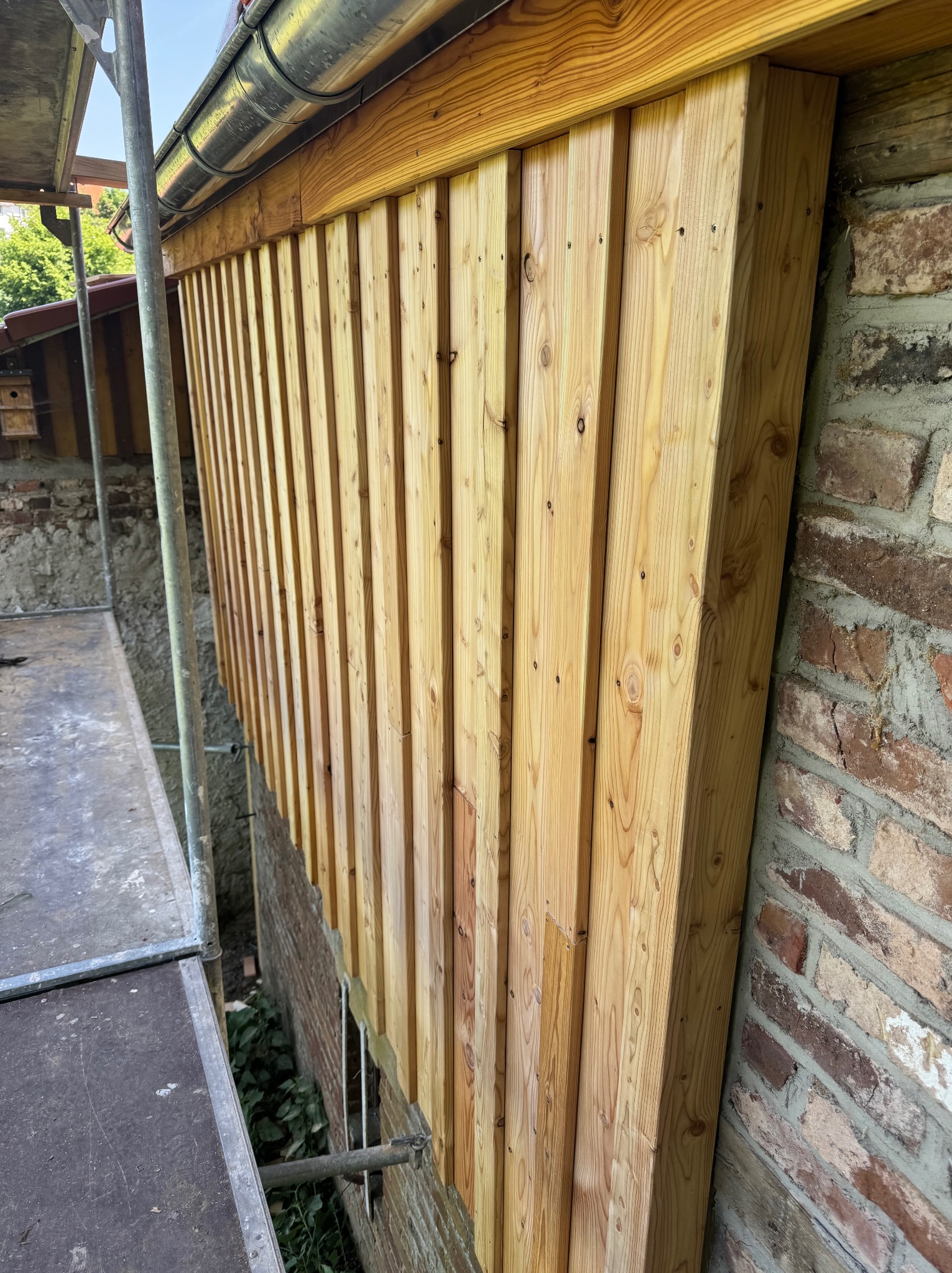 Montaż drewnianej boazerii na ścianie z cegły, widoczne rusztowanie i rynna. Boazeria z jasnego drewna, pionowe deski. Prace wykończeniowe w toku.