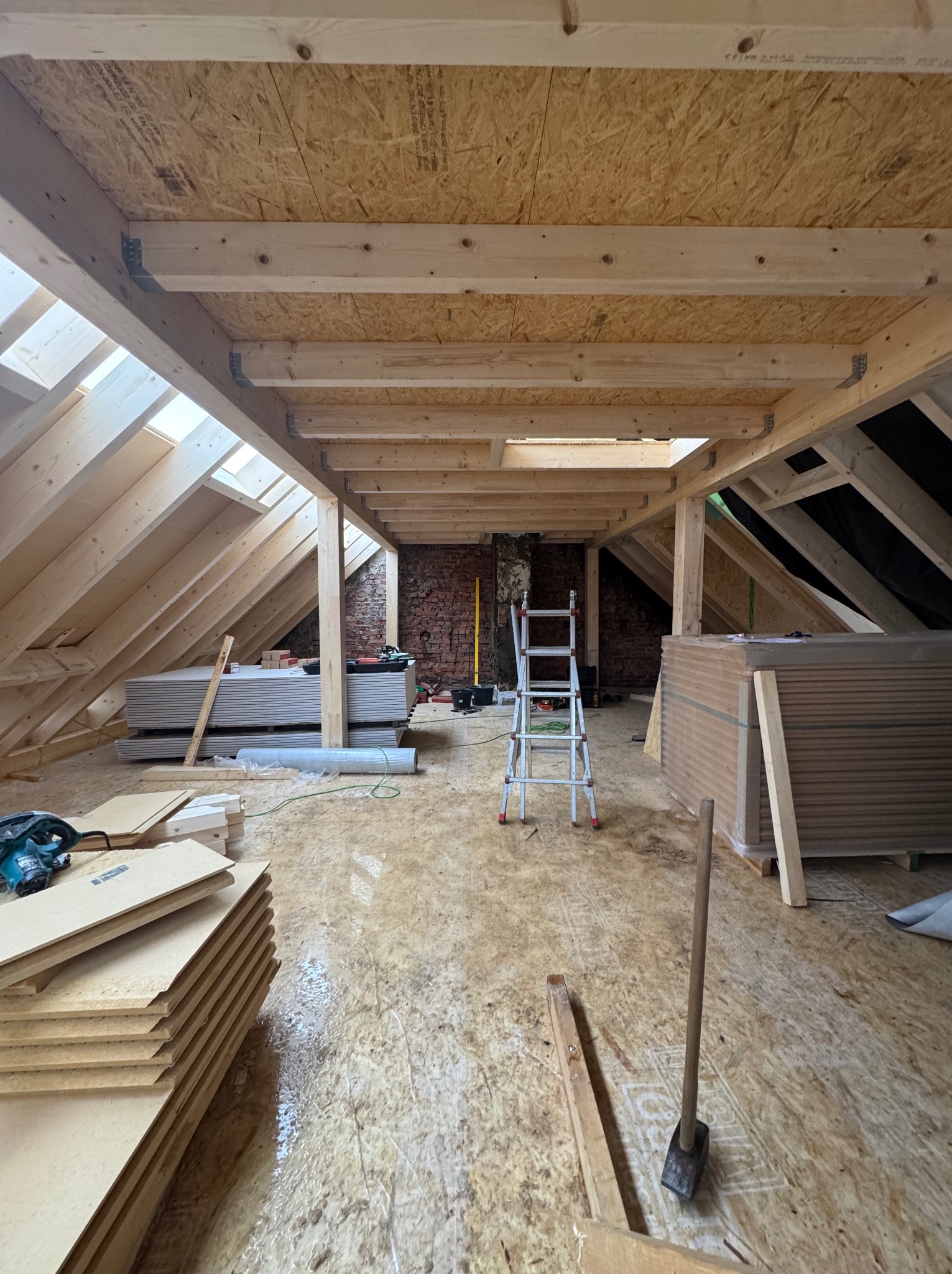 Poddasze w trakcie adaptacji z widoczną konstrukcją drewnianą, oknami dachowymi i stosem płyt kartonowo-gipsowych. W tle ceglana ściana i drabina.