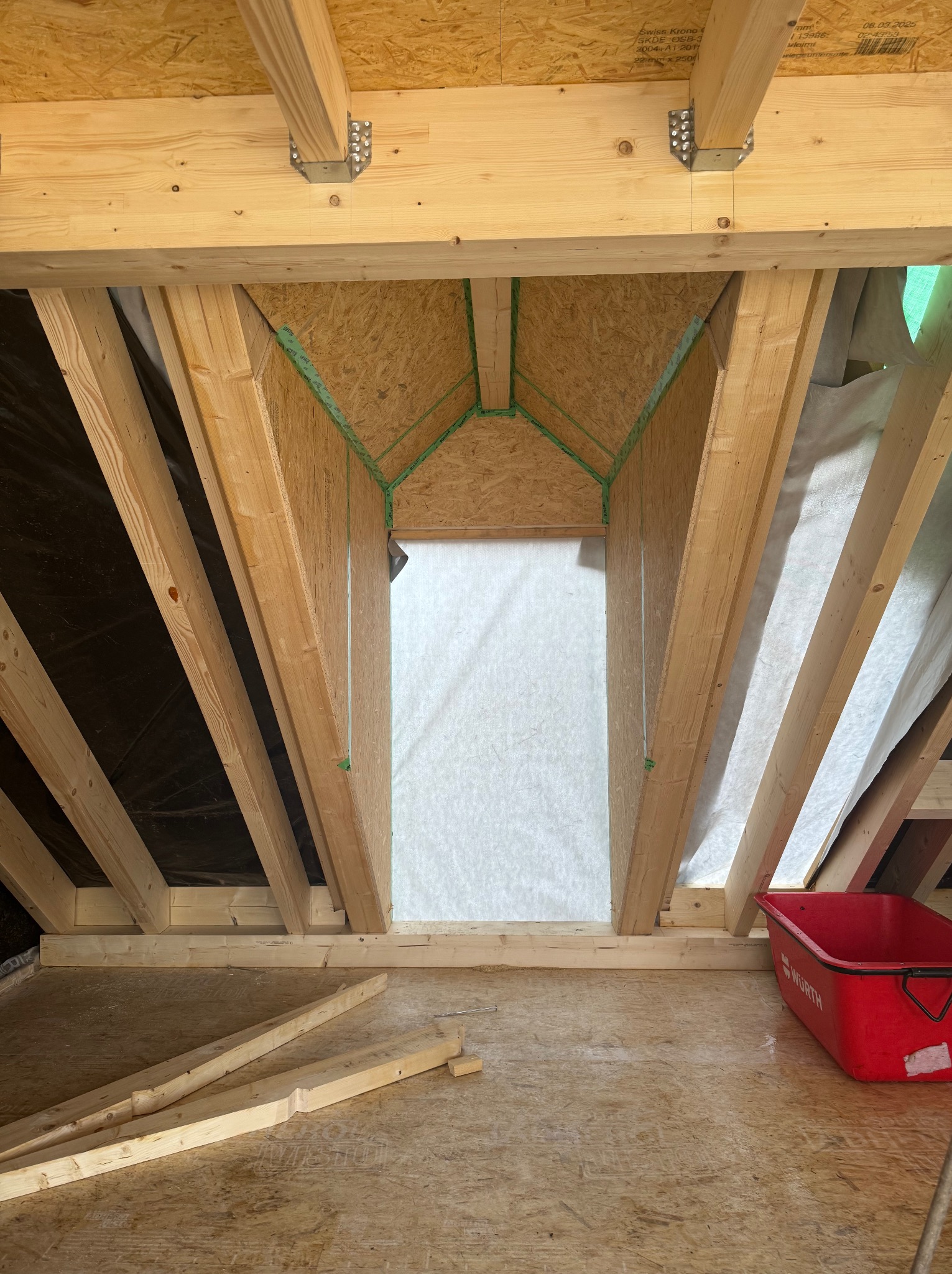 Konstrukcja drewniana poddasza z izolacją w Bobolicach, widok od wewnątrz. Elementy wzmocnione metalowymi łącznikami, widoczny fragment czerwonej taczki.