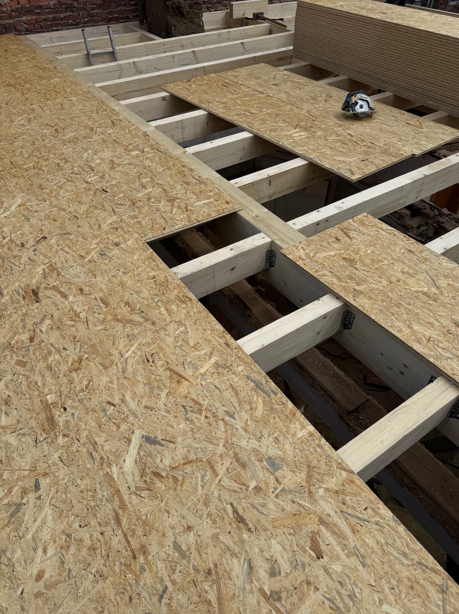 Układanie płyt OSB na drewnianej konstrukcji stropu. Widoczne elementy mocujące i narzędzie (piła) na płycie. Fragment muru w tle.