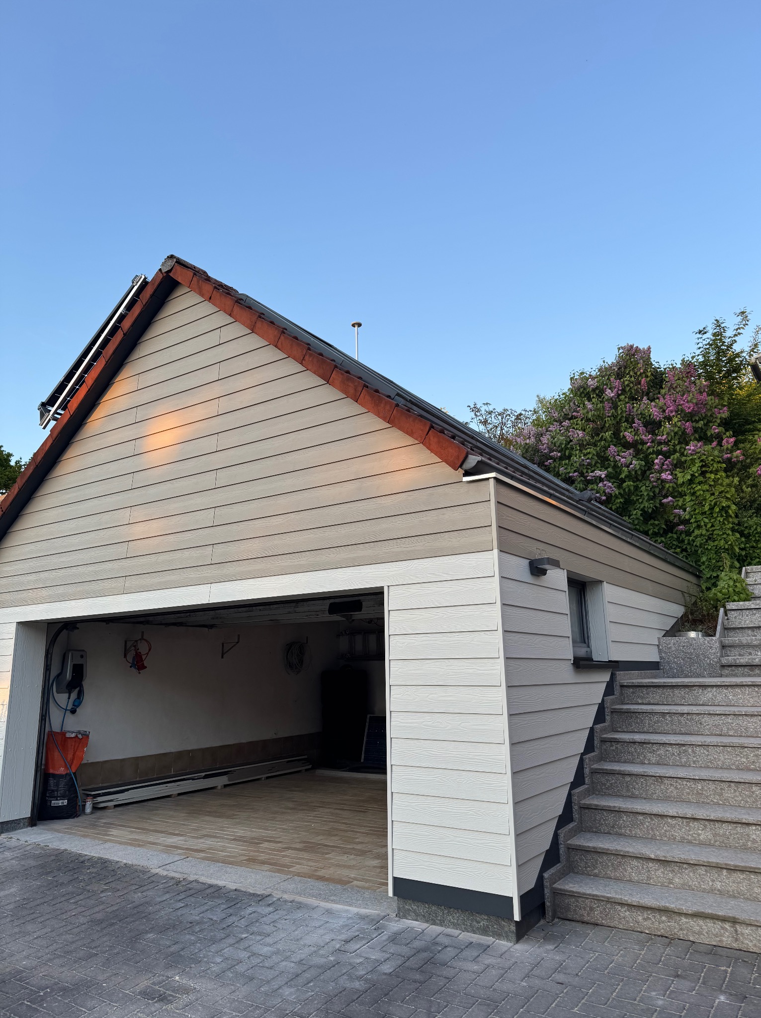 Wykończony garaż z elewacją z paneli drewnopodobnych, widoczne schody wejściowe i otwarte wrota garażowe z wnętrzem. U góry kwitnący krzew.