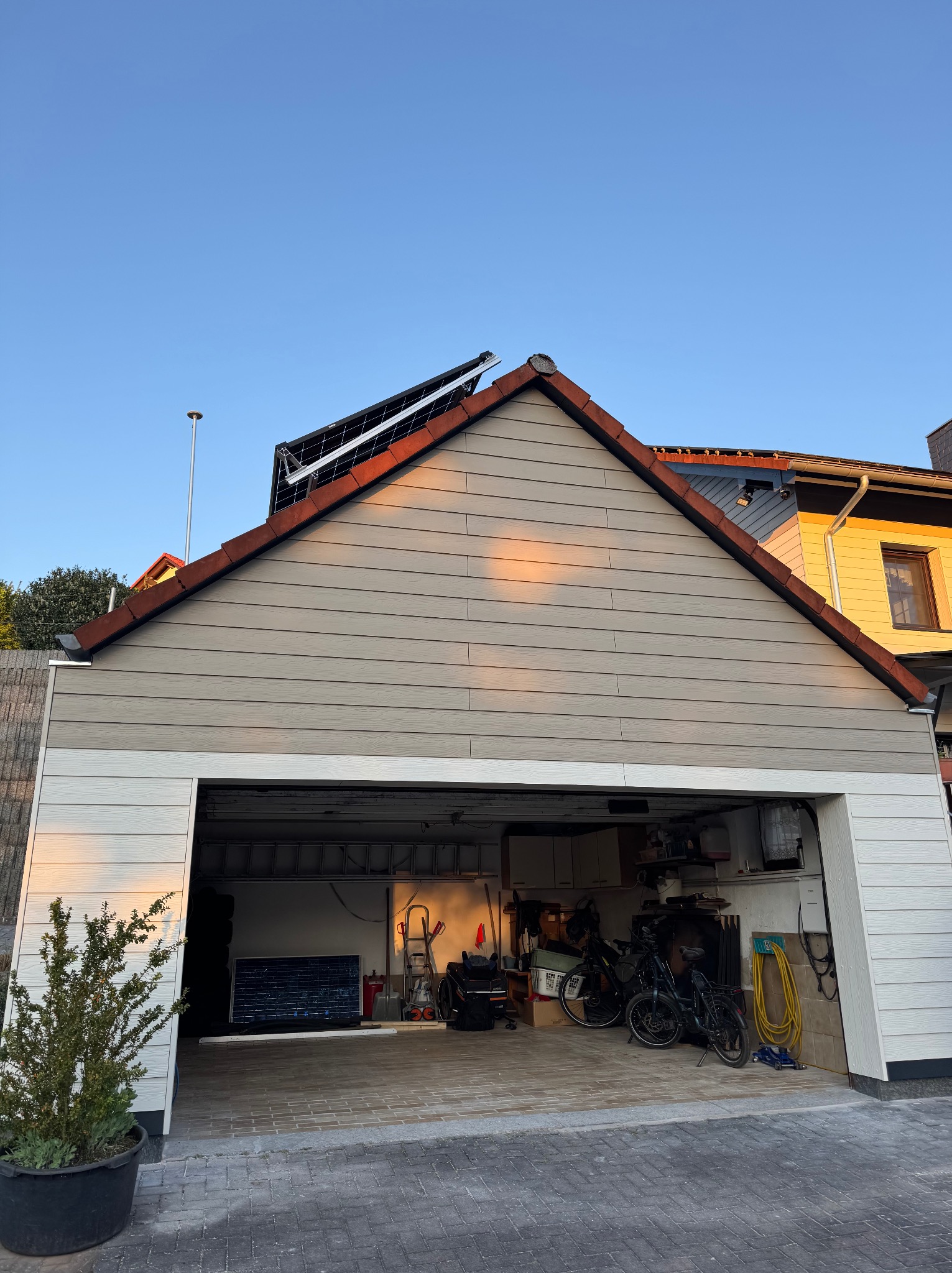 Zewnętrzna elewacja garażu z panelami w odcieniach szarości i bieli, z widocznym wnętrzem wypełnionym sprzętem. Ujęcie przy zachodzącym słońcu.