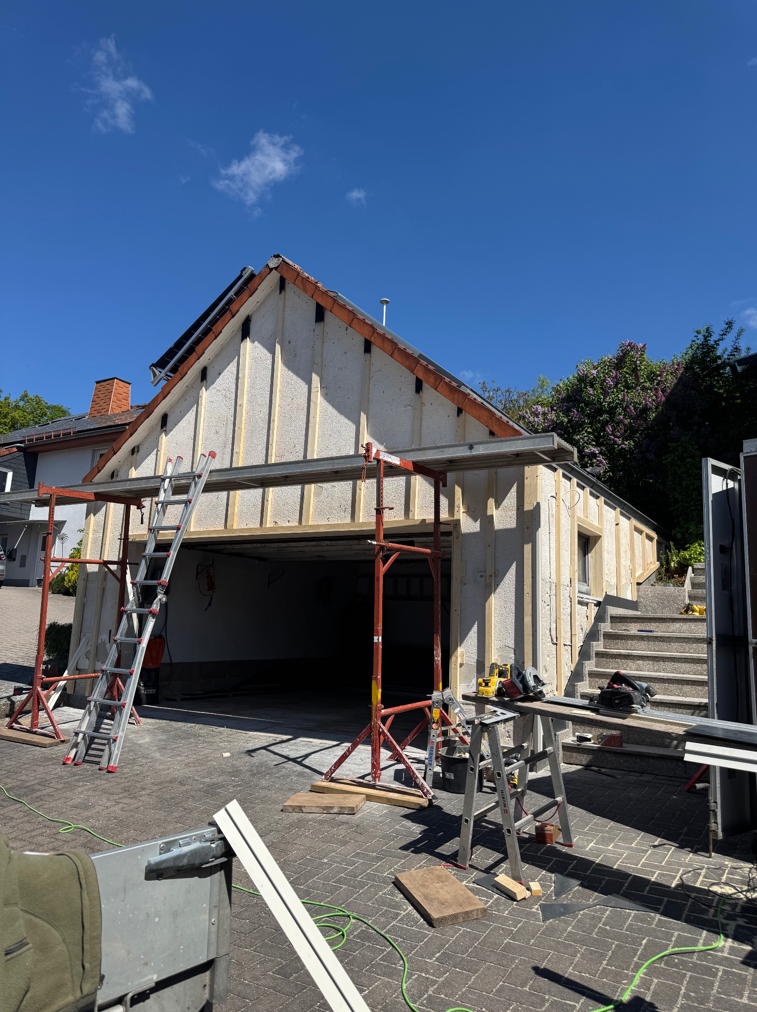 Budowa garażu z drewnianą konstrukcją szkieletową, widoczne rusztowania i narzędzia. Etap montażu ścian i dachu, jasne oświetlenie dzienne.