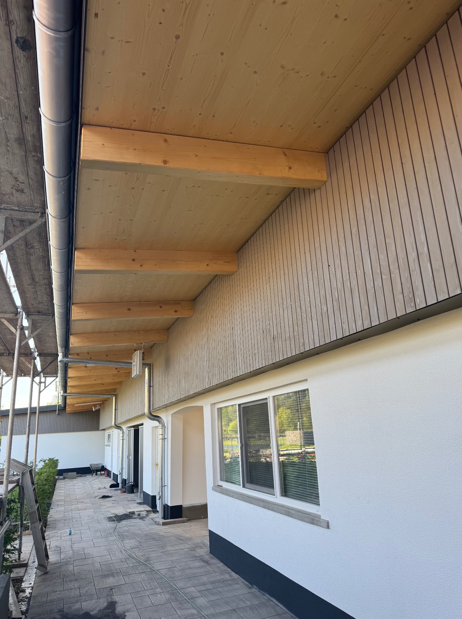 Wykończony podbitka dachowa z drewnianą boazerią i belek, elewacja budynku z oknem, rynna, chodnik z kostki brukowej, rusztowanie w tle, jasne kolory.