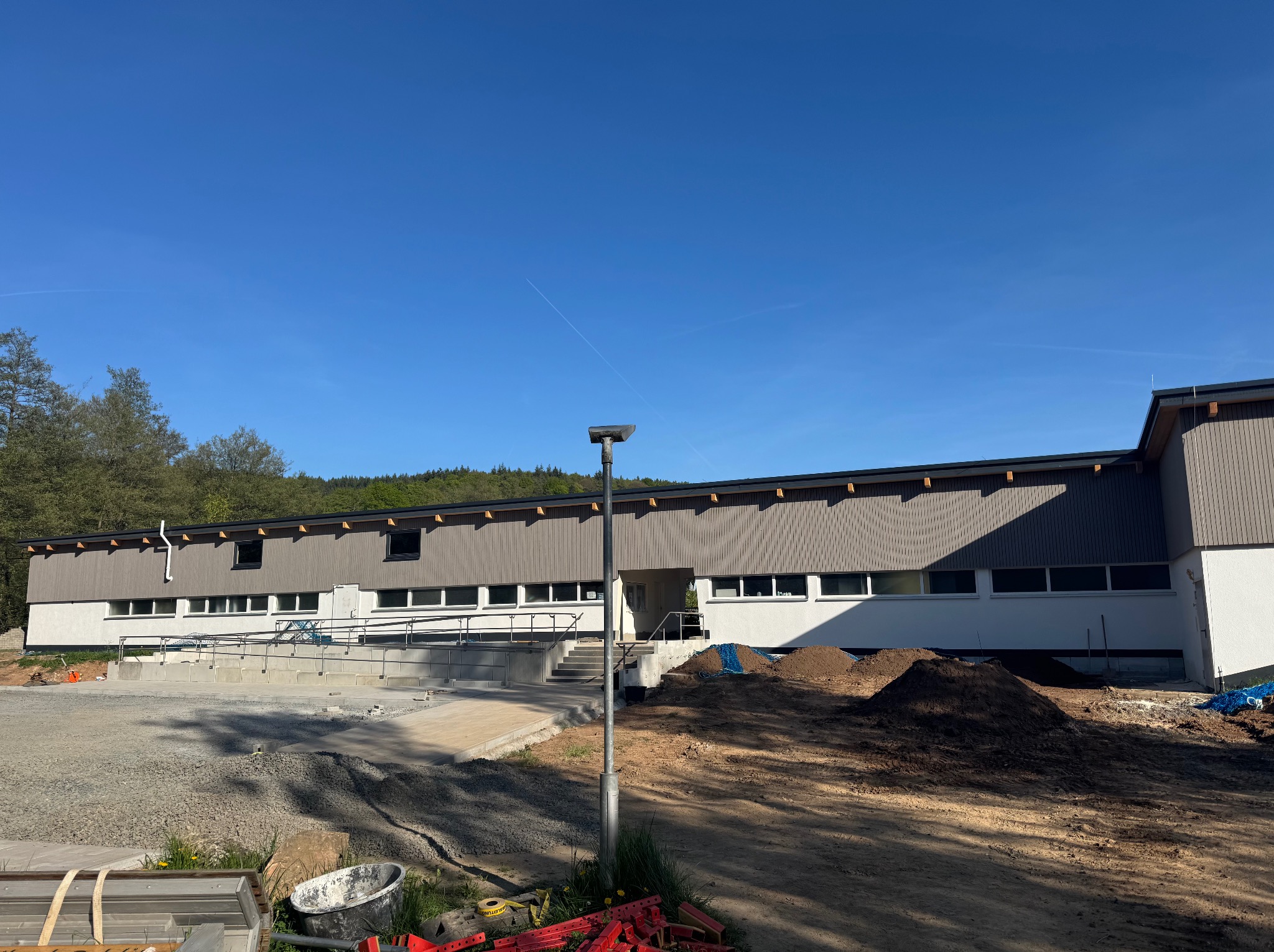 Budynek po remoncie z rampą dla niepełnosprawnych, otoczony placem budowy. Widoczne narzędzia i materiały budowlane. Jasne niebo, słoneczny dzień.