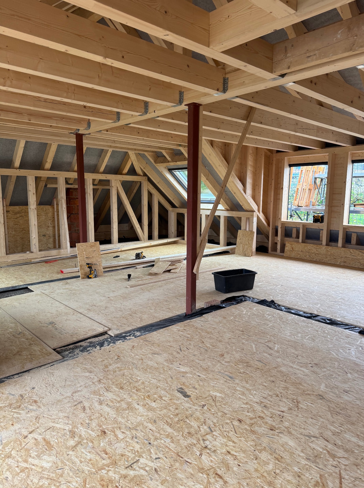 Wnętrze poddasza w trakcie adaptacji: drewniana konstrukcja szkieletowa, płyty OSB na podłodze, okna dachowe, widoczne narzędzia i materiały budowlane.