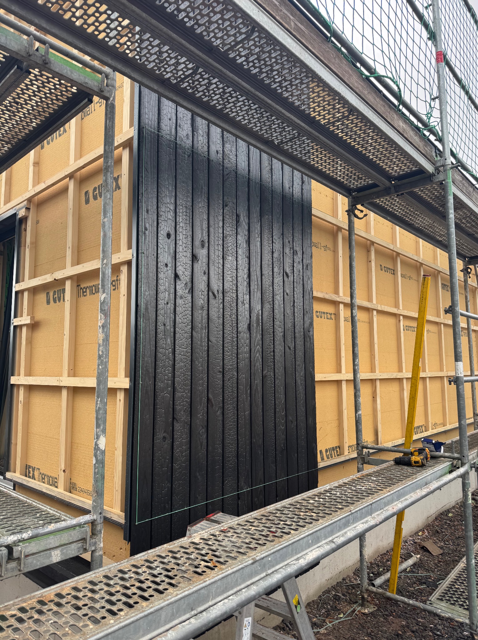 Montaż czarnej, drewnianej elewacji na budynku w trakcie budowy, widoczne rusztowania i materiały izolacyjne.