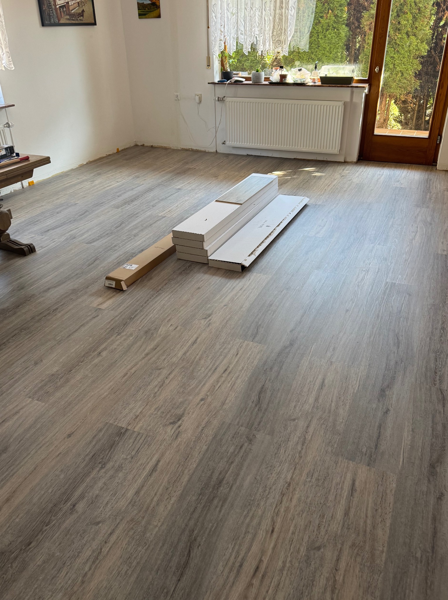 Nowa podłoga winylowa o wzorze drewna w jasnym, przestronnym wnętrzu. Częściowo rozpakowane panele czekają na montaż przy oknie z widokiem na zieleń.