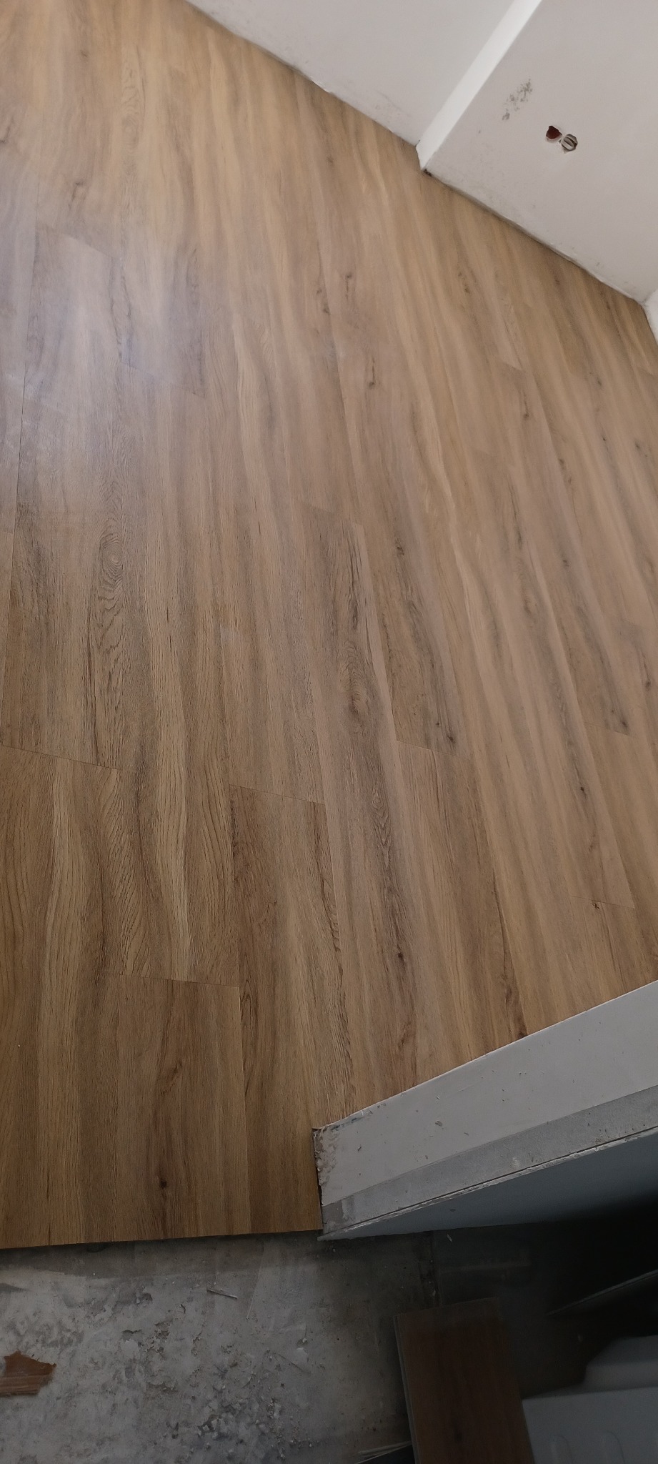 Ułożone panele podłogowe w odcieniach brązu, widoczne połączenia między panelami, fragment ściany z gniazdkiem elektrycznym oraz surowy beton w dolnej części kadru.