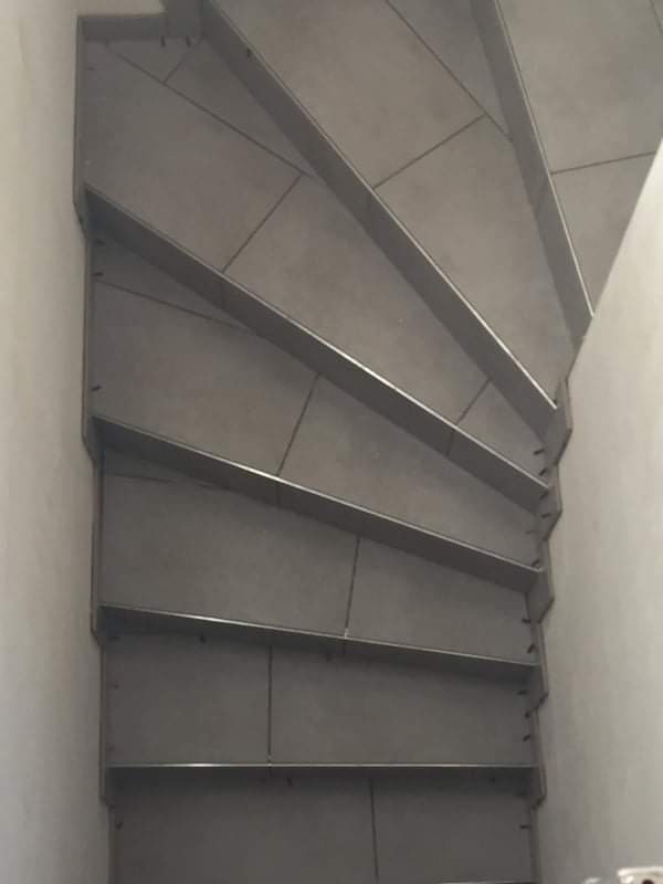 Spiralne schody wyłożone szarą, kwadratową glazurą, widok z góry, z widocznymi metalowymi krawędziami stopni.