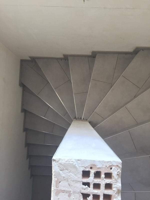 Widok z góry na spiralne schody z szarymi płytkami, z centralnym słupem o surowym wykończeniu, na tle surowego stropu.