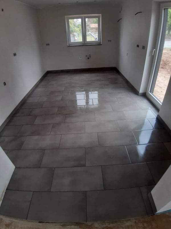 Ułożona, ciemnoszara, prostokątna terakota na podłodze w pomieszczeniu w stanie surowym; widoczne wyprowadzenia instalacji na ścianach, okno z widokiem na zewnątrz.