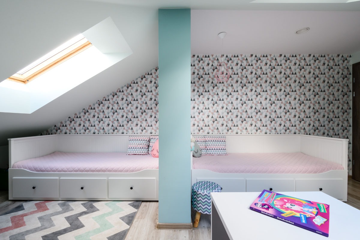 Pokój dziecięcy na poddaszu z dwoma białymi łóżkami, geometryczną tapetą i oknem dachowym. Pastelowa kolorystyka, dywan w zygzaki i stolik z książką.
