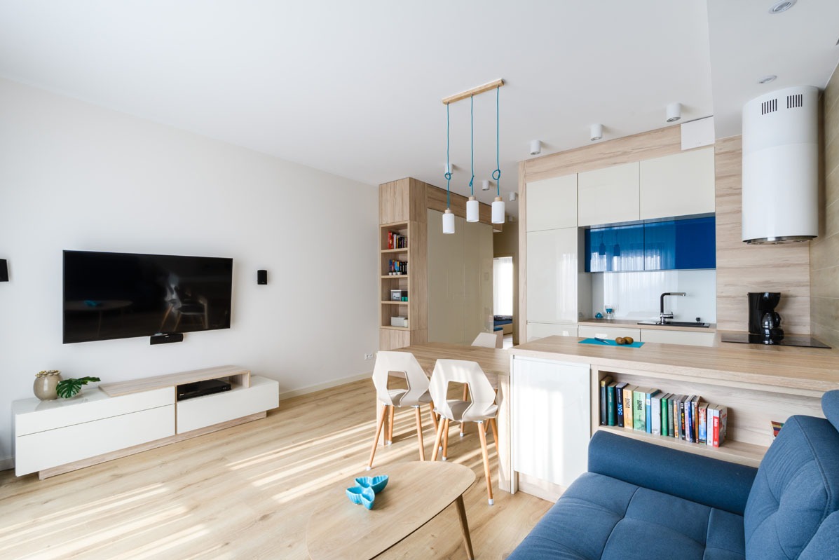 Jasne, nowoczesne wnętrze: salon z aneksem kuchennym. Białe ściany, drewniana podłoga, niebieska sofa. Minimalistyczny design, oświetlenie punktowe i wiszące lampy.