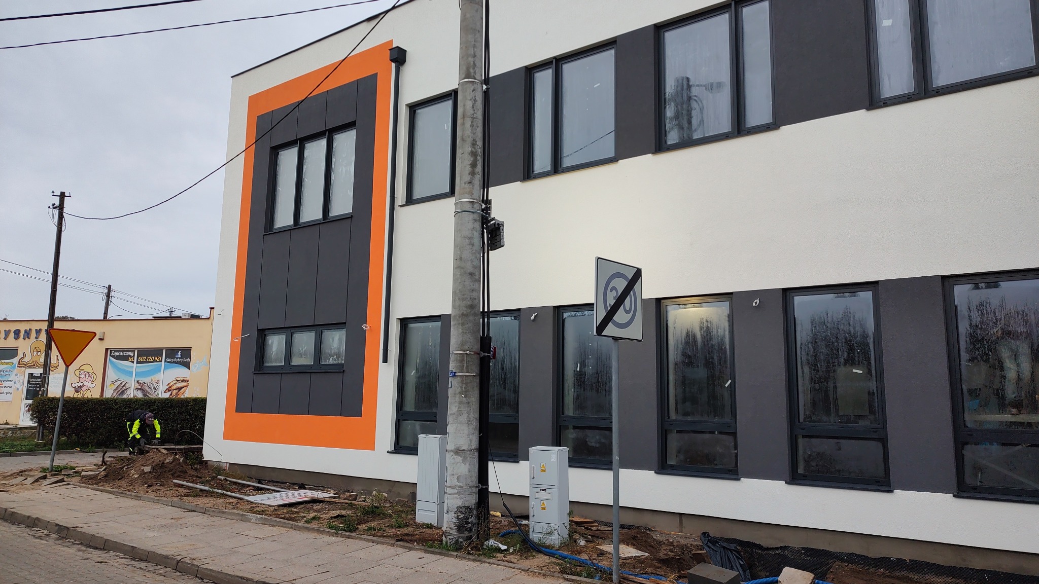 Nowoczesny budynek z elewacją w odcieniach bieli i szarości, z pomarańczowym akcentem. Widoczne okna z czarnymi ramami i słup energetyczny. Prace ziemne w toku.