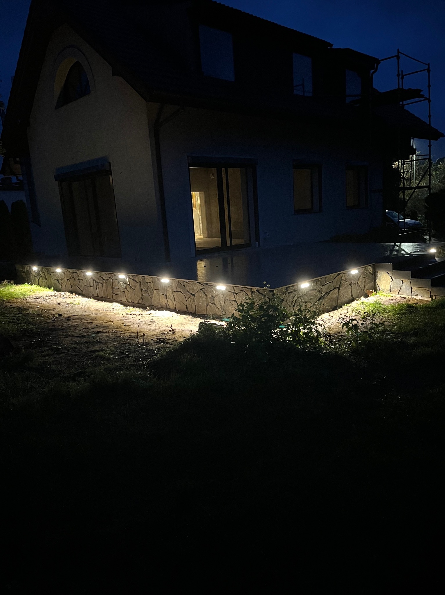 Nocne oświetlenie elewacji domu z kamienną podmurówką, z lampami wzdłuż ścieżki i na schodach. Widoczne rusztowanie przy budynku.