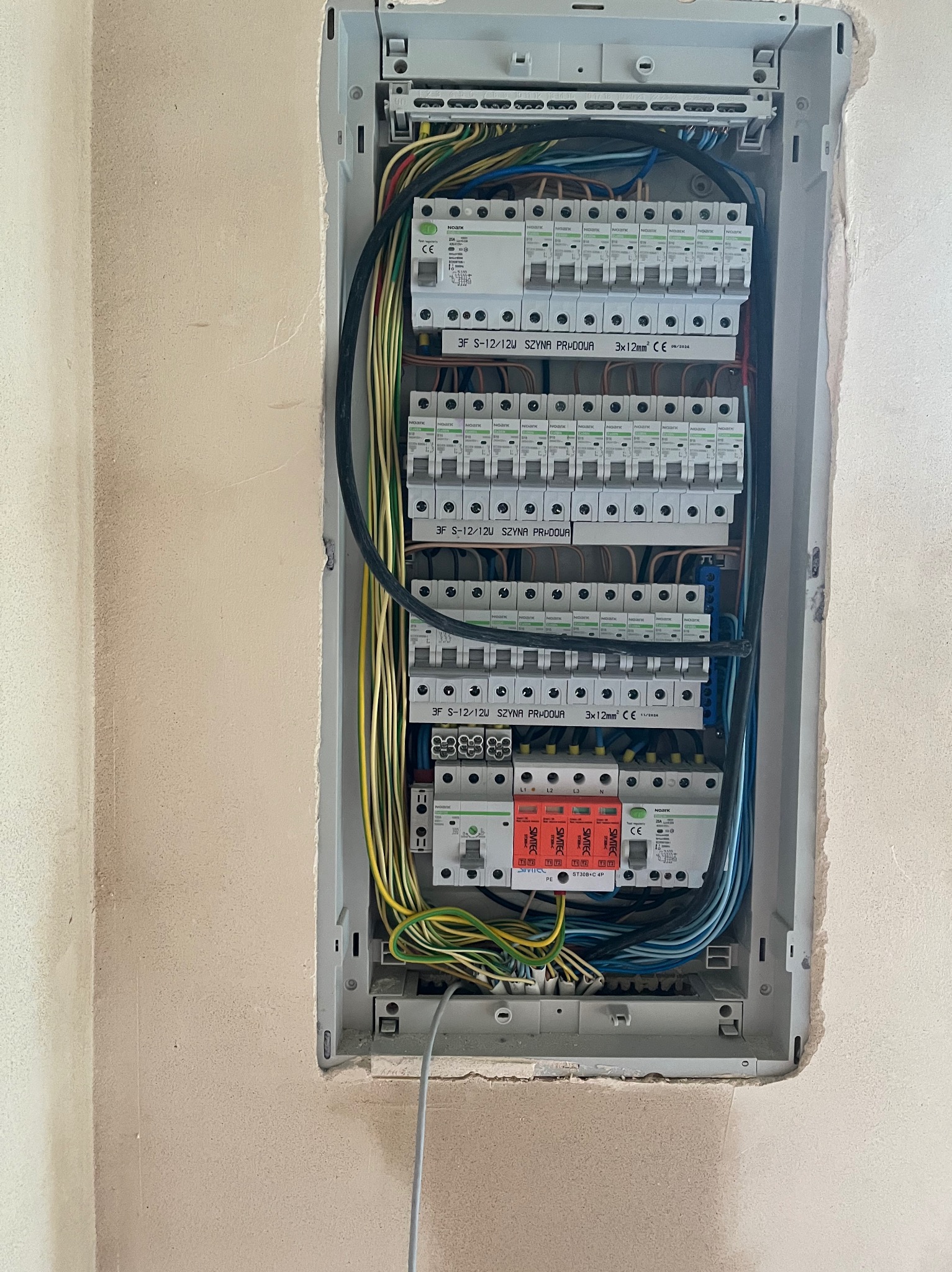 Rozdzielnica elektryczna z zainstalowanymi bezpiecznikami i okablowaniem, wnęka w ścianie, widoczne przewody w kolorach żółtym, zielonym, niebieskim i czarnym.