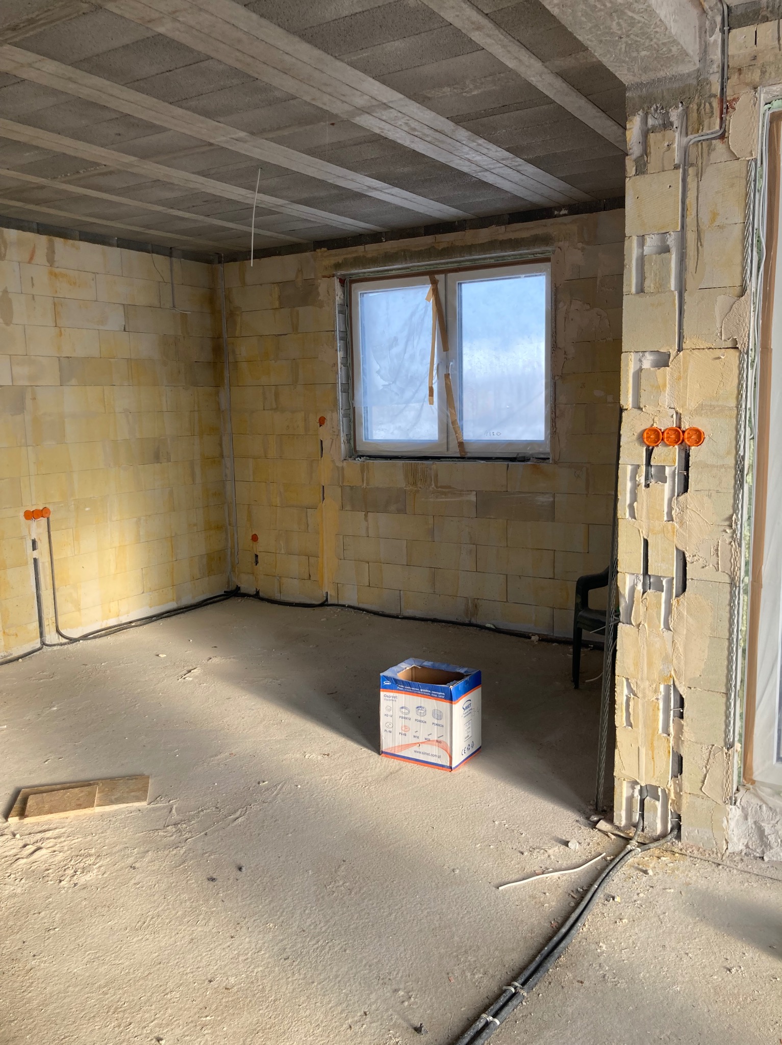 Instalacja elektryczna w surowym wnętrzu: widoczne kable w ścianach z betonu komórkowego, puszki elektryczne i skrzynka na podłodze. Okno w tle.