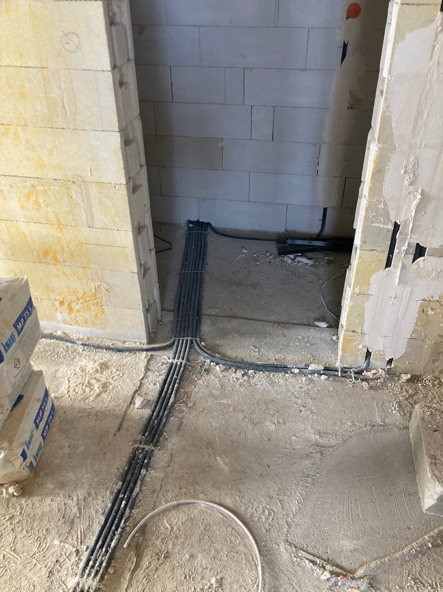 Wiązka czarnych kabli elektrycznych w peszlu ułożona na podłodze w surowym stanie budynku z bloków betonu komórkowego. Instalacja elektryczna w trakcie prac.