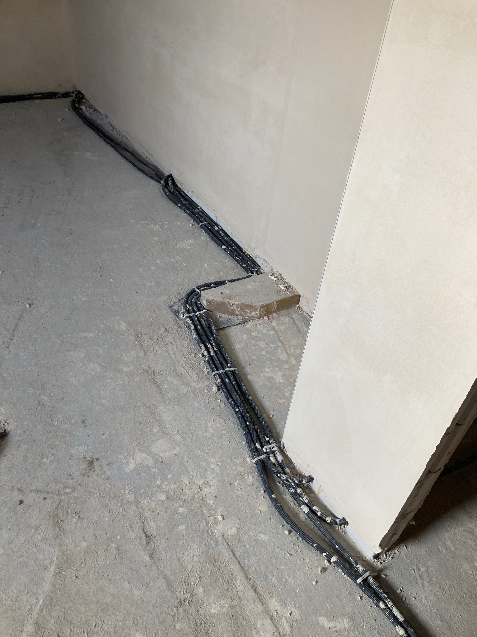 Szczegółowe ujęcie przewodów elektrycznych w peszlach, prowadzonych wzdłuż ściany na surowym, betonowym podłożu wewnątrz budynku w stanie deweloperskim.