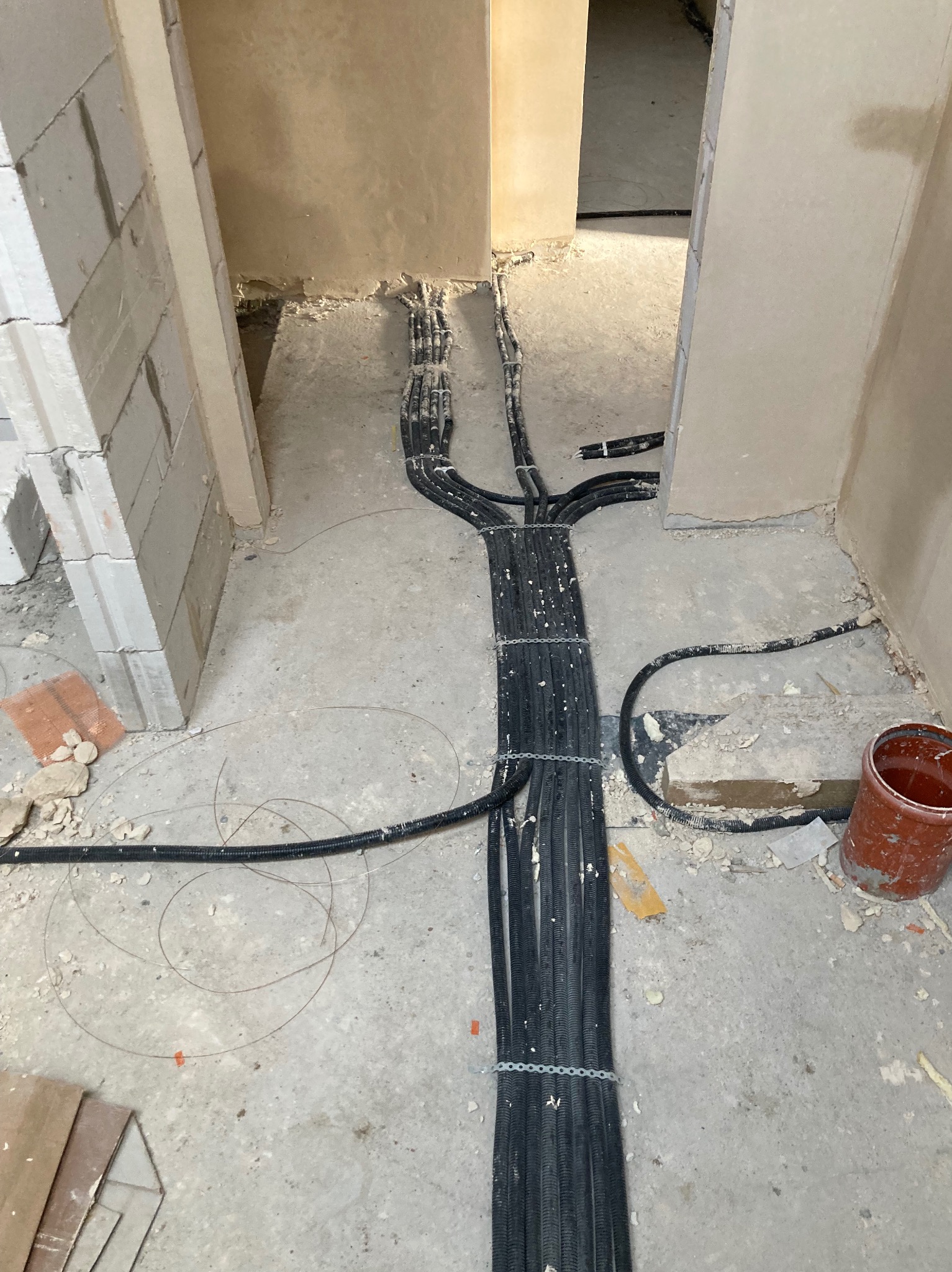 Rozprowadzone kable w peszlach, częściowo przymocowane do podłoża, w surowym wnętrzu w trakcie prac wykończeniowych. Widok z góry, kable rozchodzą się promieniście.