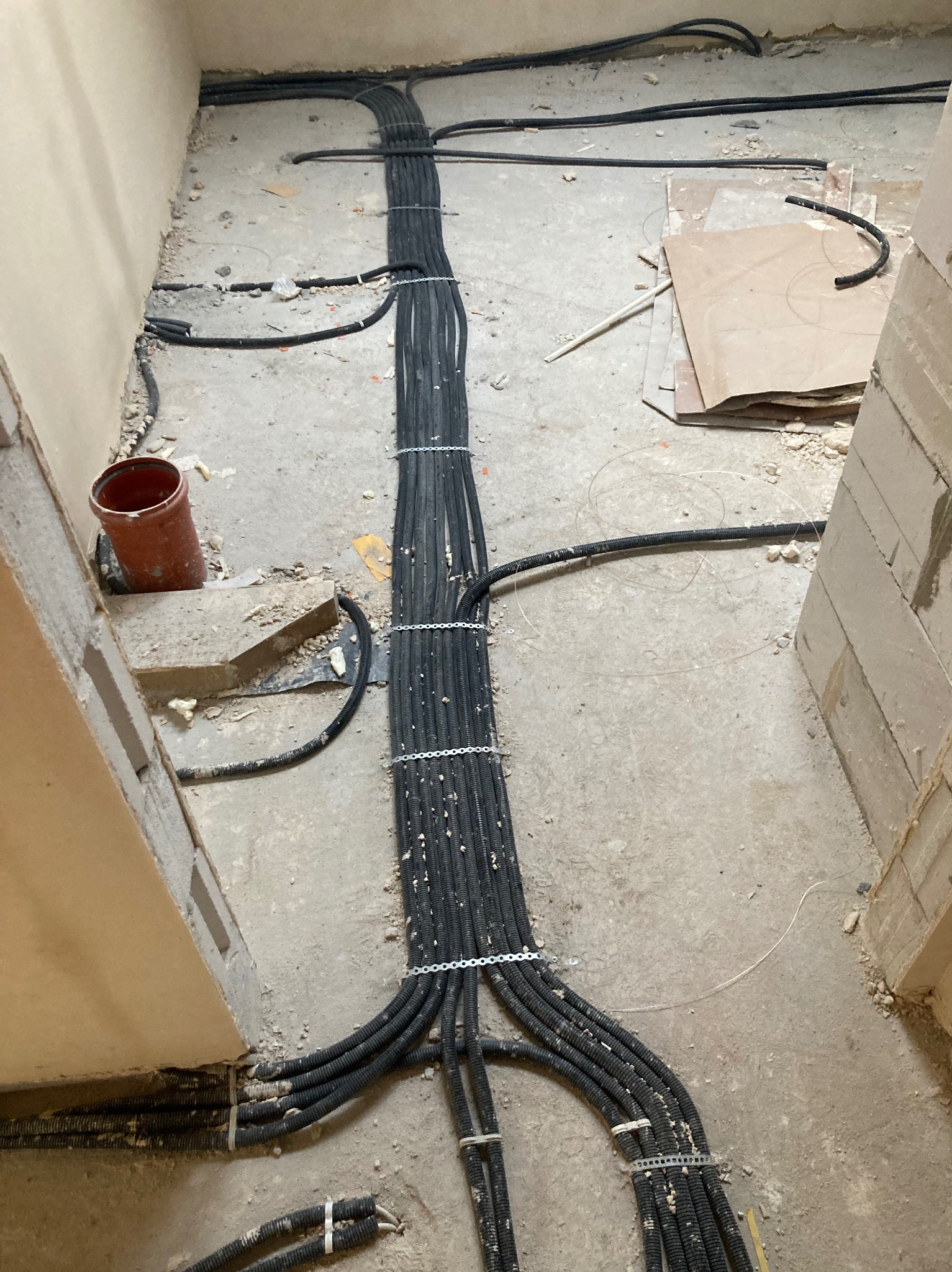 Wiązka czarnych kabli w peszlach, przymocowanych metalowymi uchwytami, ułożona na surowej podłodze w trakcie prac instalacyjnych wewnątrz budynku.