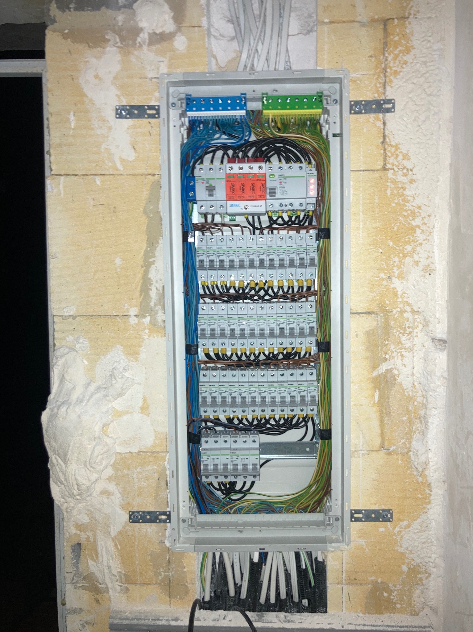 Rozdzielnica elektryczna w trakcie montażu, widoczne liczne przewody, bezpieczniki i elementy montażowe. Ściana w surowym stanie, z widocznymi śladami prac murarskich.