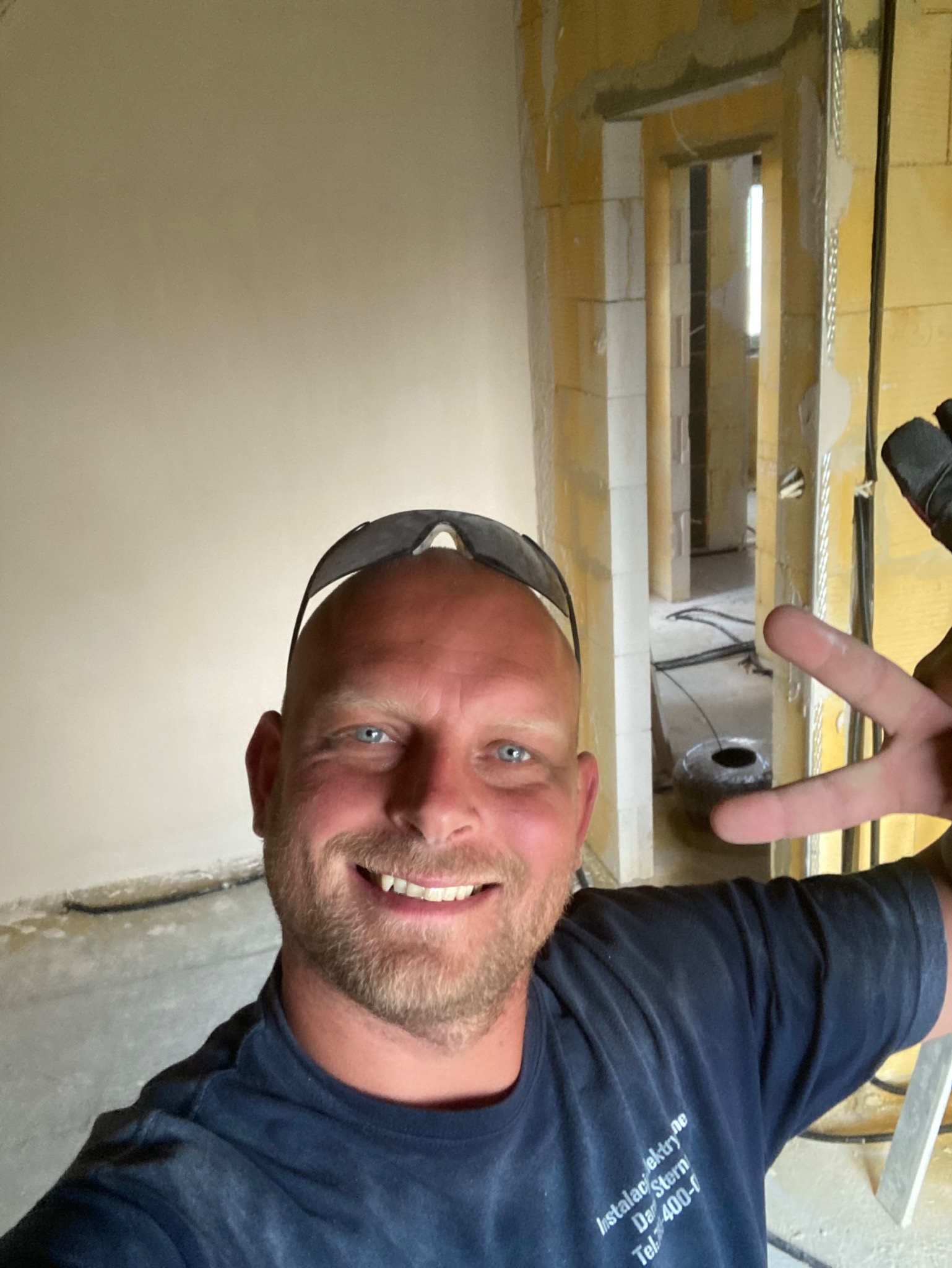 Elektryk w trakcie prac instalacyjnych, uśmiechnięty robi selfie na tle surowego wnętrza z widocznymi przewodami i otworami. W tle widoczne kolejne pomieszczenia.