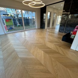 GRAND INTERIORS SPÓŁKA Z OGRANICZONĄ ODPOWIEDZIALNOŚCIĄ - Jasny parkiet ułożony we wzór jodełki w przestronnym salonie z dużym oknem i nowoczesnym oświetleniem. Widoczny fragment czarnej ściany z marmurowym wzorem.