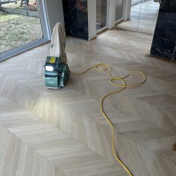 GRAND INTERIORS SPÓŁKA Z OGRANICZONĄ ODPOWIEDZIALNOŚCIĄ - Maszyna do renowacji parkietu na jasnej podłodze ułożonej w jodełkę, z widocznym przewodem zasilającym. W tle okno i fragment ściany z czarnym marmurem.