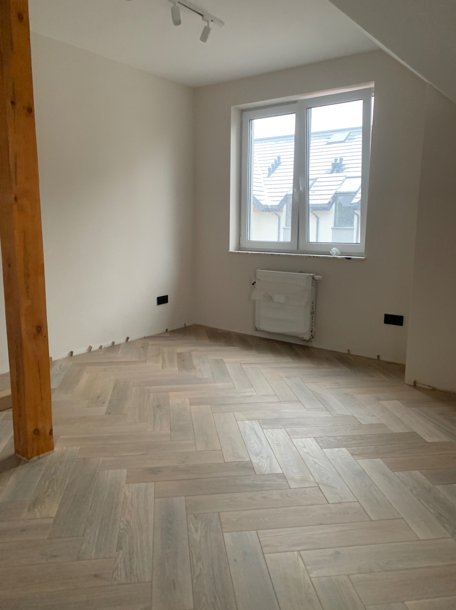 Jasne parkiet ułożony we wzór jodełki w pokoju z oknem i drewnianym słupem. Grzejnik zabezpieczony folią. Widoczne listwy przypodłogowe i gniazdka elektryczne.