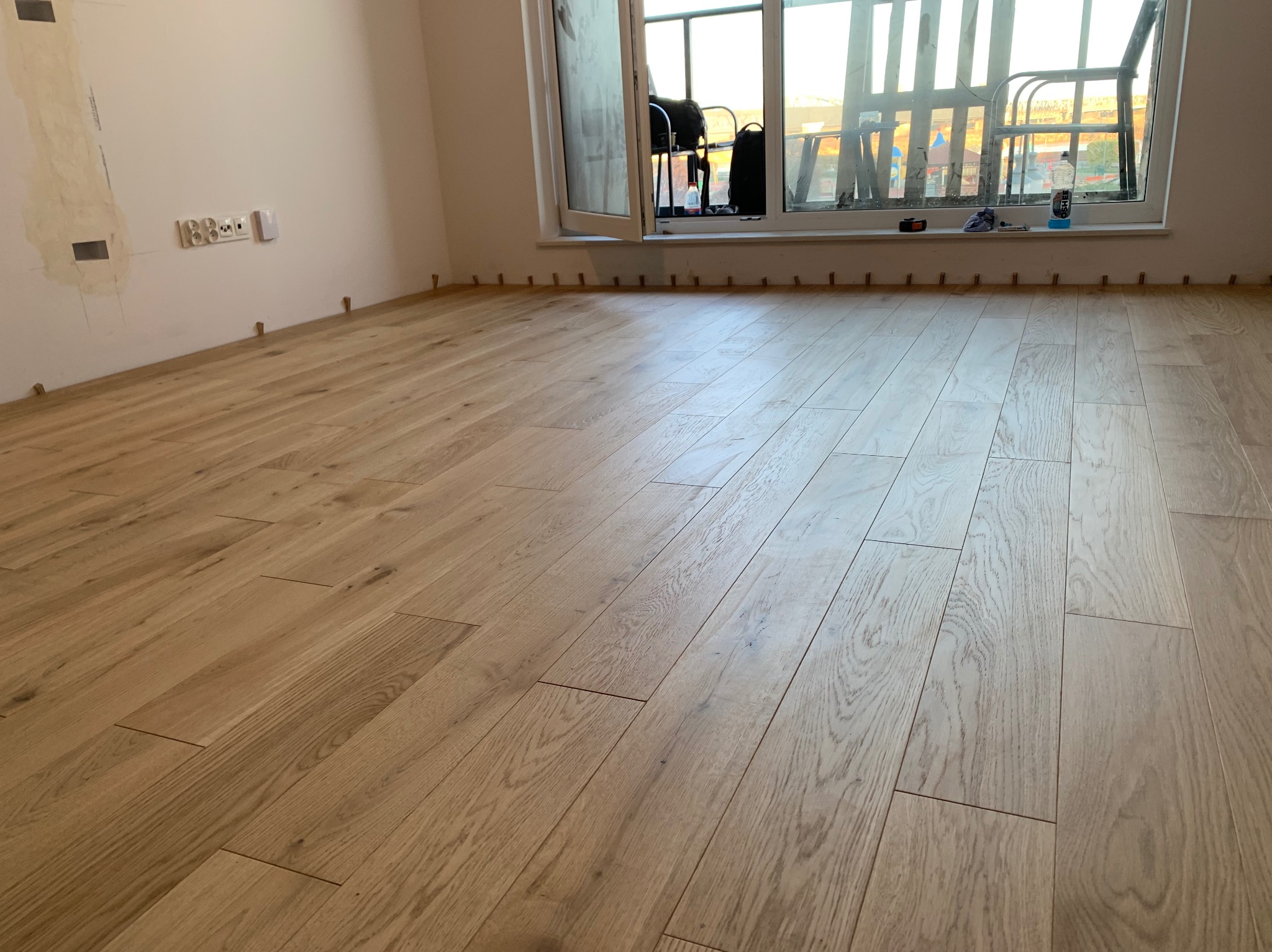 Jasna, drewniana podłoga ułożona w pokoju z oknem balkonowym. Widoczne listwy dystansowe przy ścianach i gniazdka elektryczne. Naturalne światło.