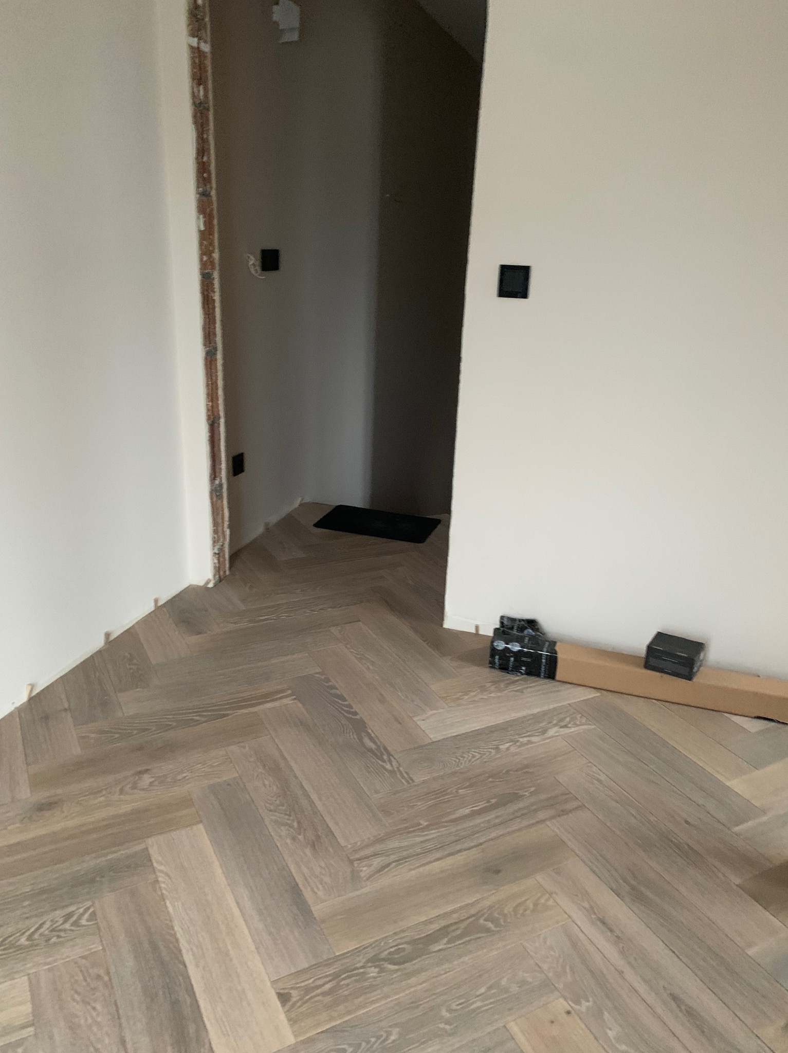 Ułożony parkiet w jodełkę francuską, widoczne paczki paneli przy ścianie. Realizacja z Warszawy, parkiet w odcieniach szarości i beżu, surowe ściany.