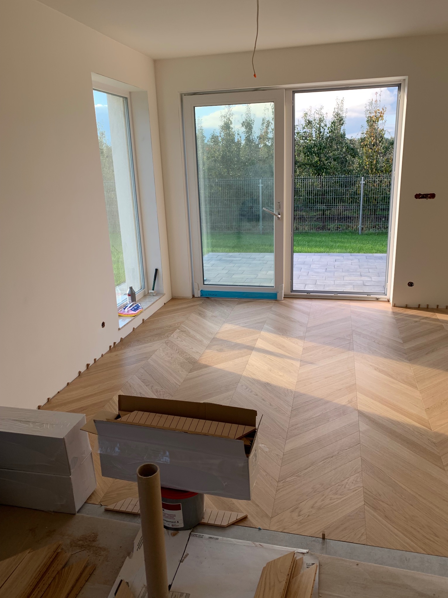 Jasne parkiety ułożone we wzór jodełki francuskiej w nowym wnętrzu z dużym oknem balkonowym. Widoczne materiały i narzędzia podczas montażu.