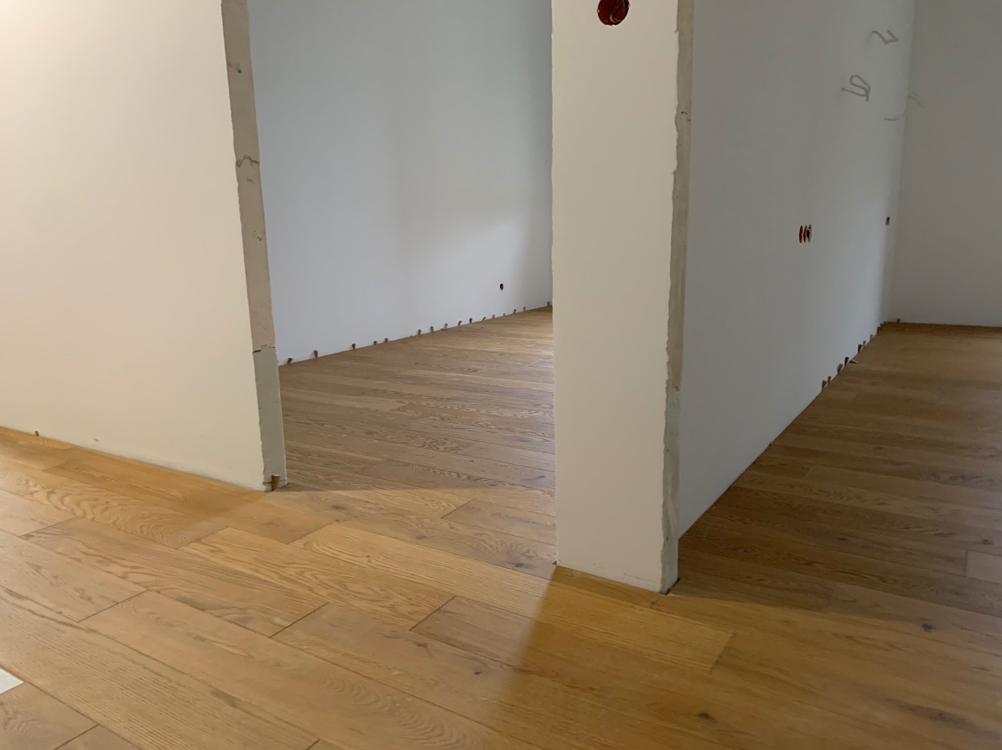 Jasne, drewniane deski podłogowe w nowym wnętrzu, widok przez surowy otwór drzwiowy. Fragmenty ścian z instalacją elektryczną. Kołki dystansowe przy ścianach.