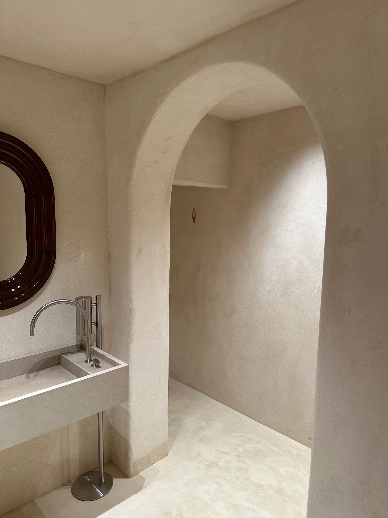 Beżowa łazienka z umywalką na podeście, okrągłym lustrem w ciemnej ramie i zaokrąglonym przejściem do kolejnego pomieszczenia. Minimalistyczny design.