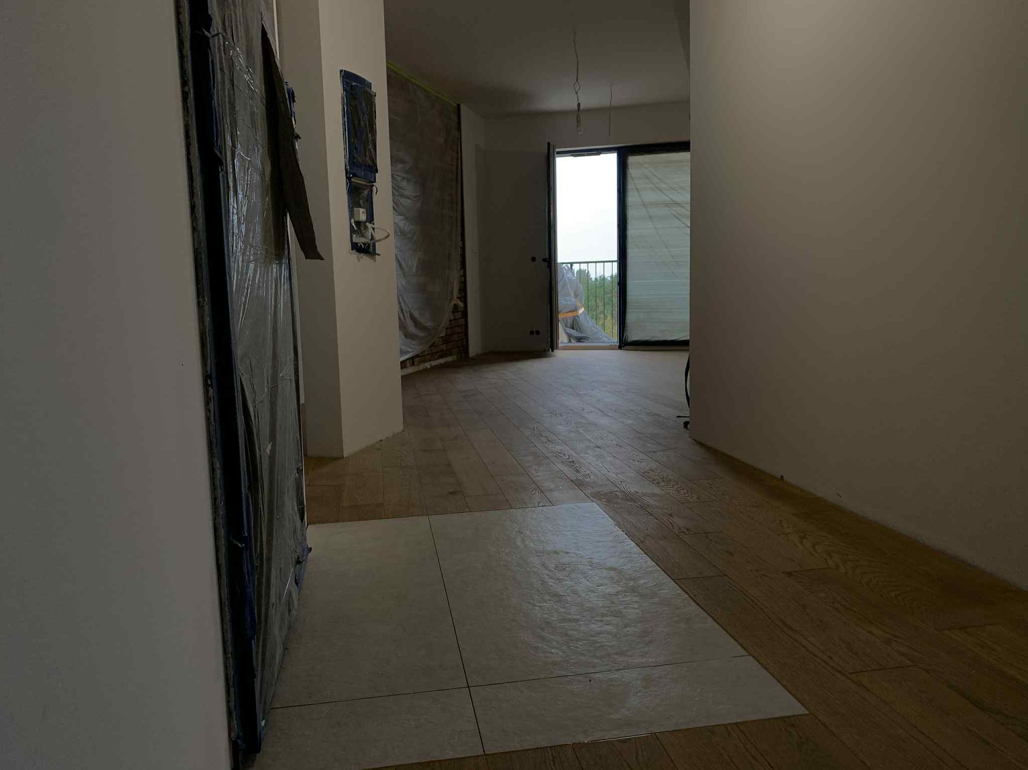 Wnętrze w trakcie prac. Fragment ułożonej podłogi drewnianej i płytek. Ściany przygotowane do malowania, okna balkonowe z widokiem na zieleń.