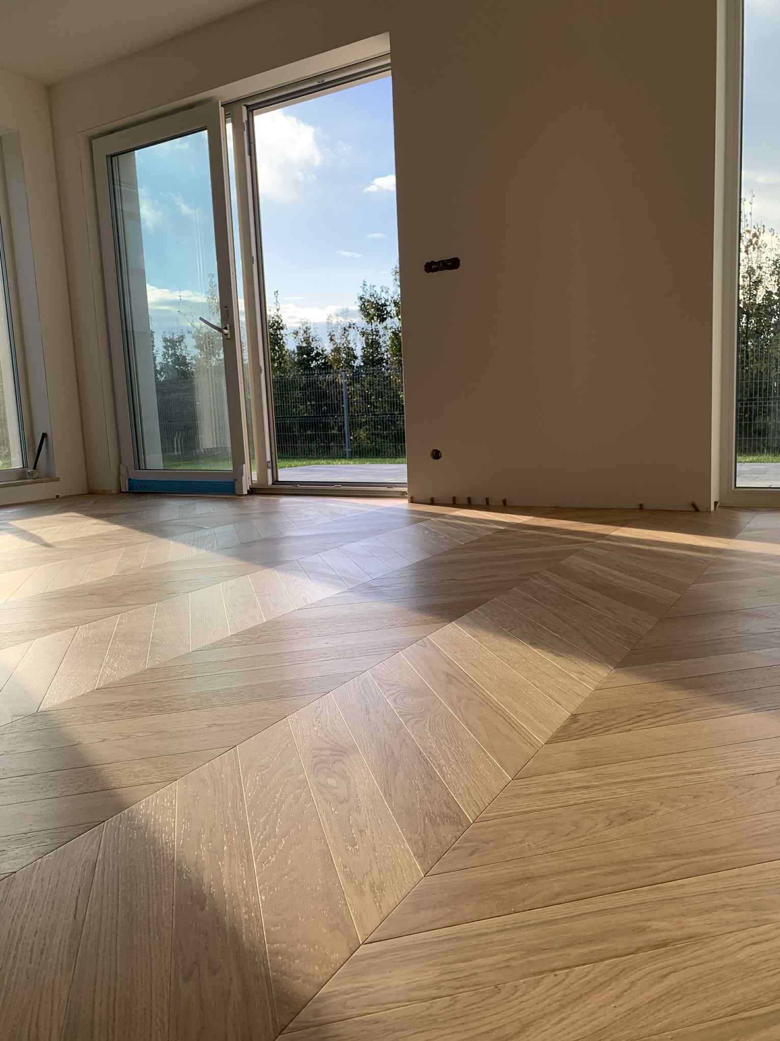 Jasne parkiet ułożony we wzór jodełki francuskiej, widok na taras przez otwarte drzwi balkonowe. Naturalne oświetlenie podkreśla strukturę drewna.