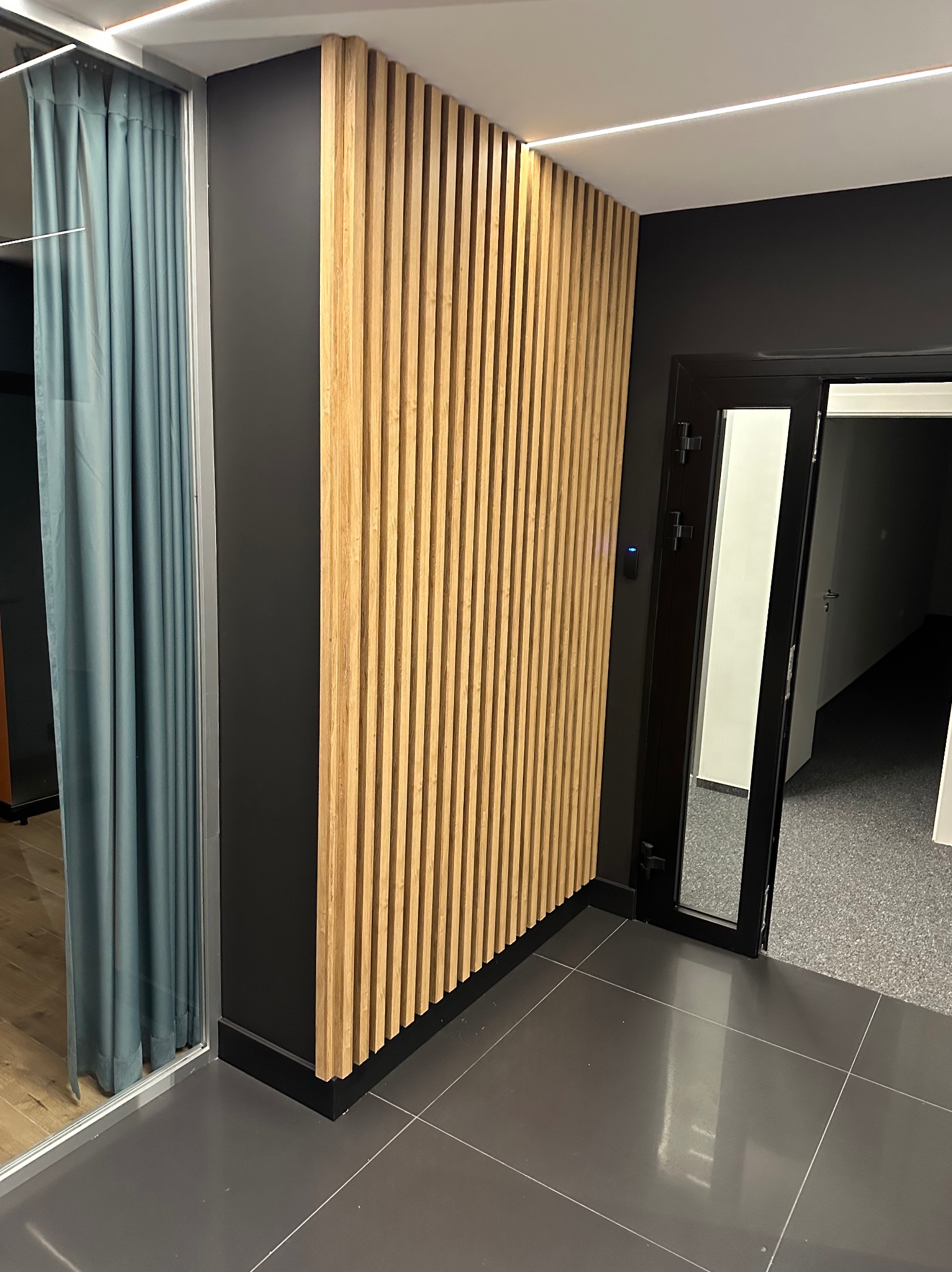 Nowoczesne wnętrze z drewnianą ścianą lamelową, szarą podłogą i czarnymi elementami wykończenia. Widoczne drzwi i zasłonięte okno.