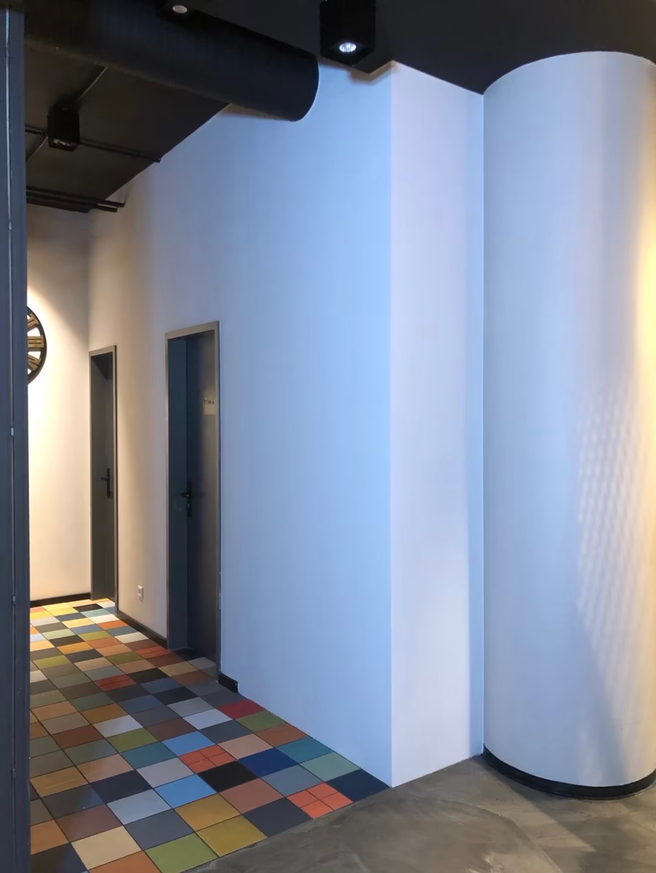 Korytarz biurowy z kolorową podłogą z kwadratowych płytek, białe ściany i dwie pary drzwi. Widoczny fragment zegara na ścianie.