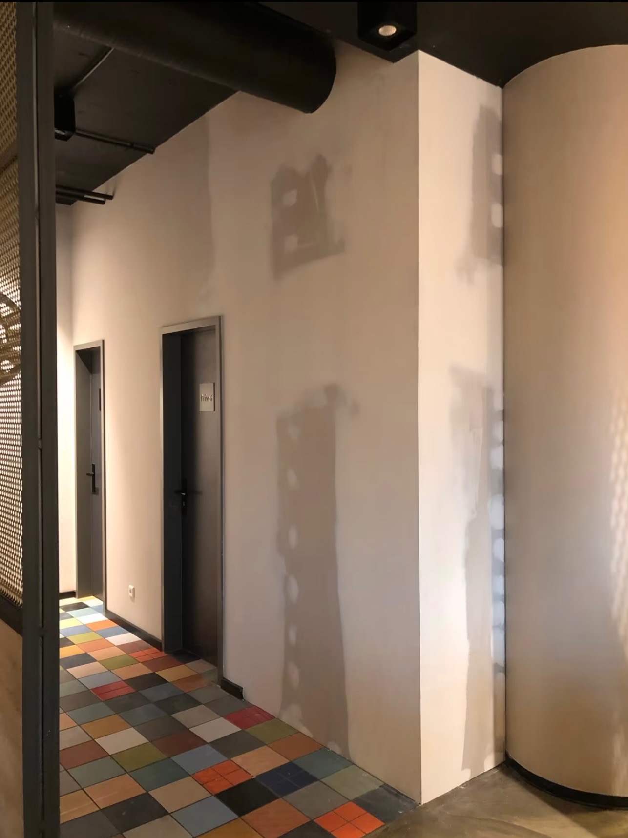 Korytarz w trakcie prac wykończeniowych: ściany z widocznymi śladami szpachlowania, kolorowa podłoga z płytek, dwoje drzwi z szarymi ościeżnicami.